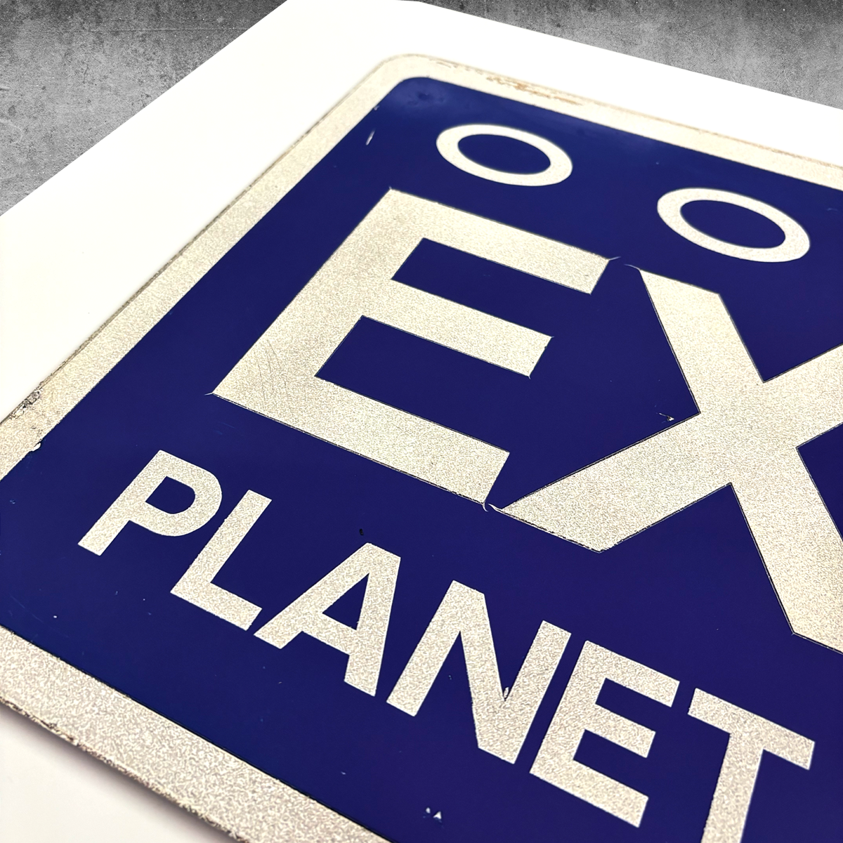 A close-up of the RocknRoad Sign Print - Exit Planet Dust by My Store, a limited edition of 25, featuring EXPLANET with two stylized eyes above EX, metallic reflective border, and a white background.