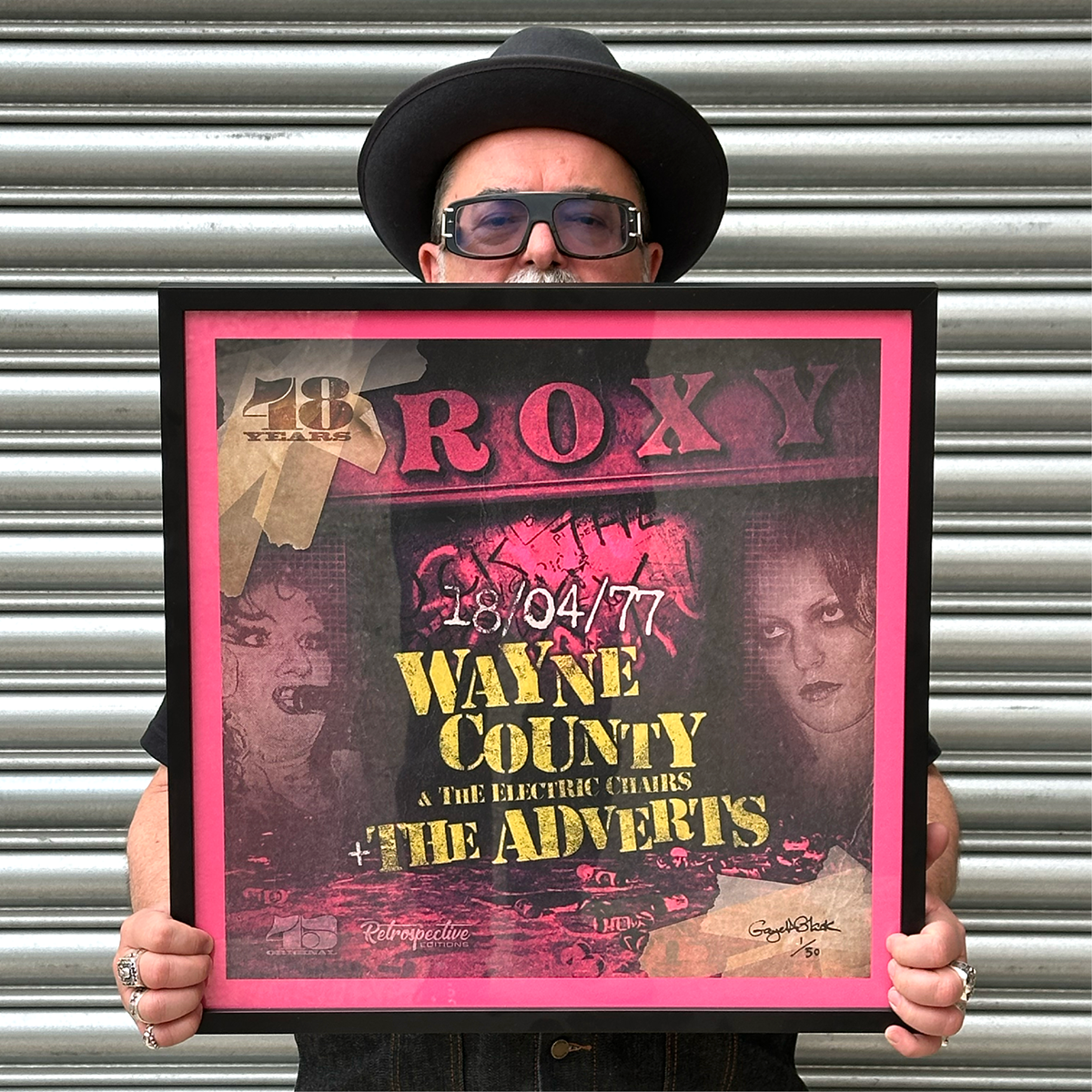 A person in a black hat and glasses holds a framed 45 Original The Adverts / Wayne County - Roxy Club On This Night Limited Edition Poster Print (signed by Gaye Black), standing in front of a metal shutter.