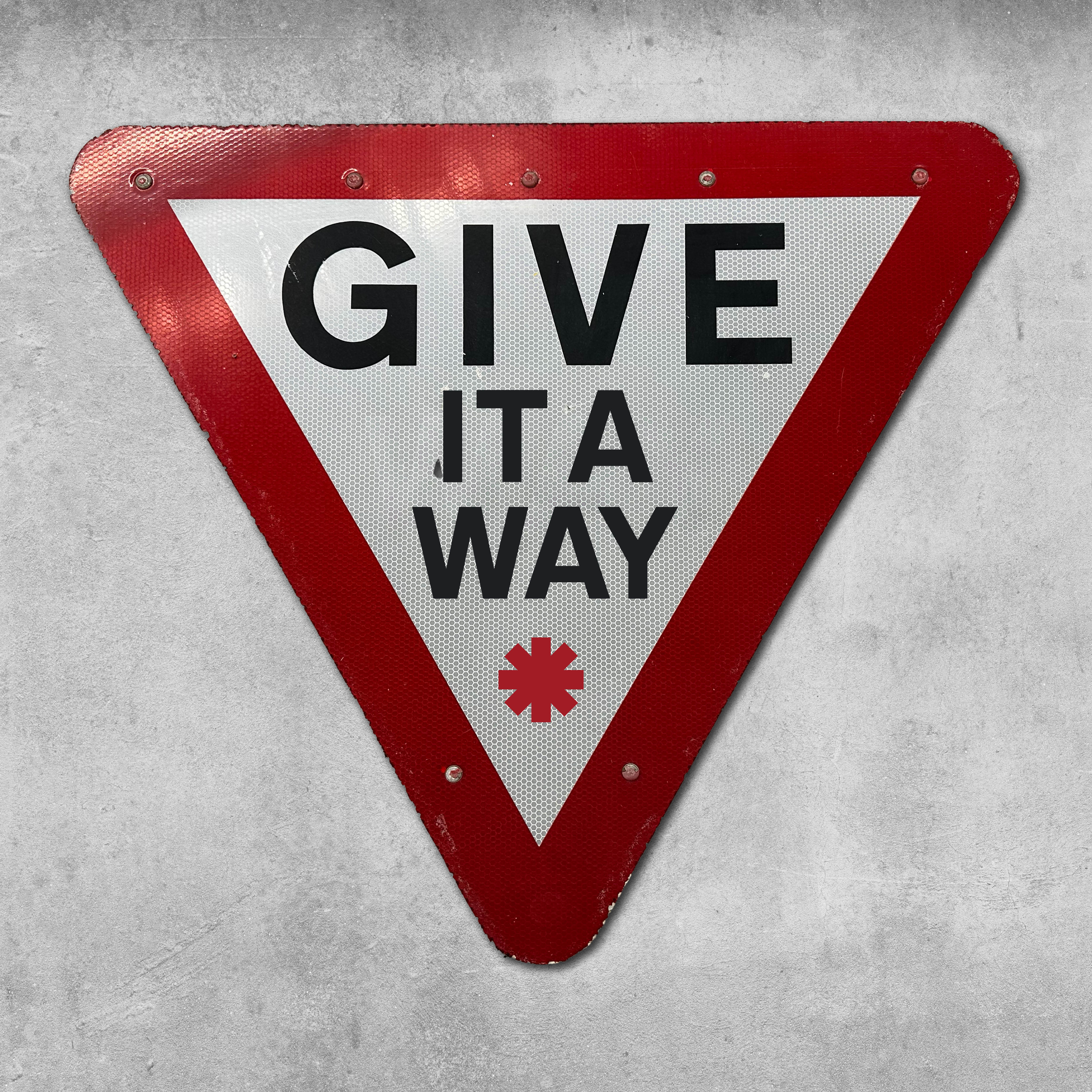 The 45 Original RocknRoad Sign - Give It A Way is a triangular UK metal road sign with a red border, white center, bold black text, and red asterisk. It has a reflective surface and is mounted on a gray concrete wall.