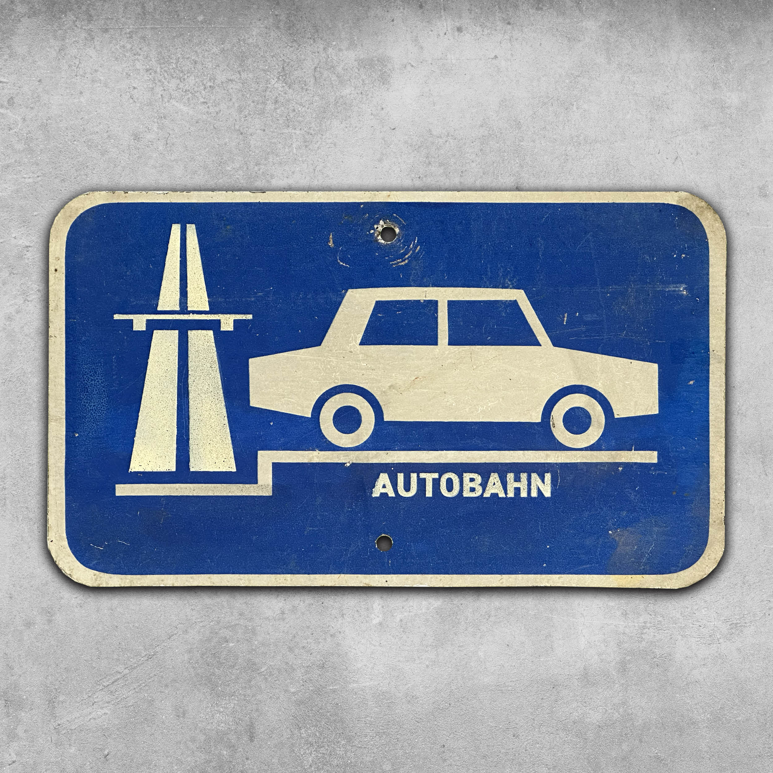 The Original Rock’n’Road Sign - Autobahn by My Store is a vintage-style blue and white East German road sign with a Trabant car and highway icon, labeled AUTOBAHN, and weathered patina, mounted on concrete.
