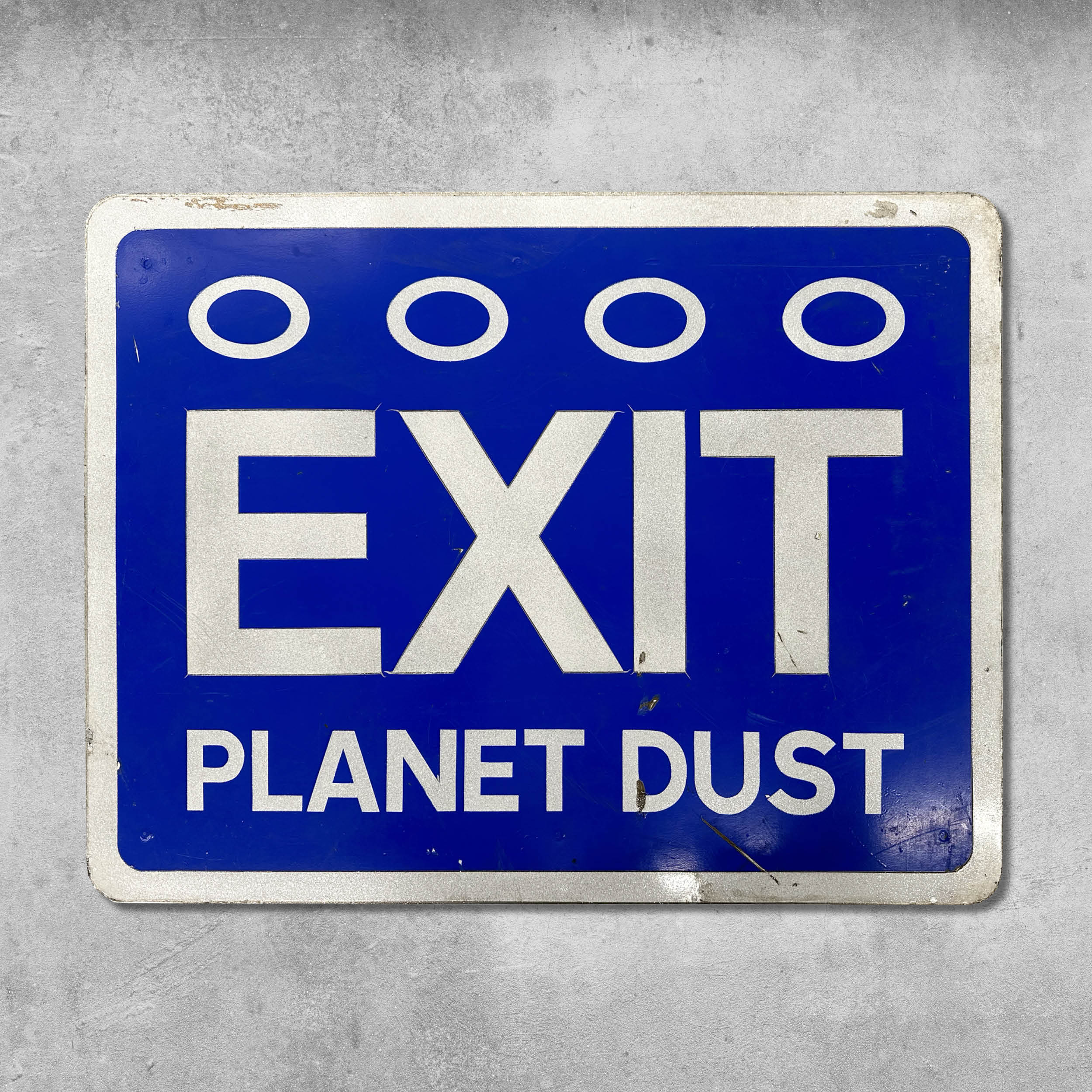 The Original RocknRoad Sign - Exit Planet Dust by My Store features bold blue and white lettering, four white circles above EXIT, a subtle sparkle finish, and is mounted on a gray concrete wall—an homage to an iconic dance album.