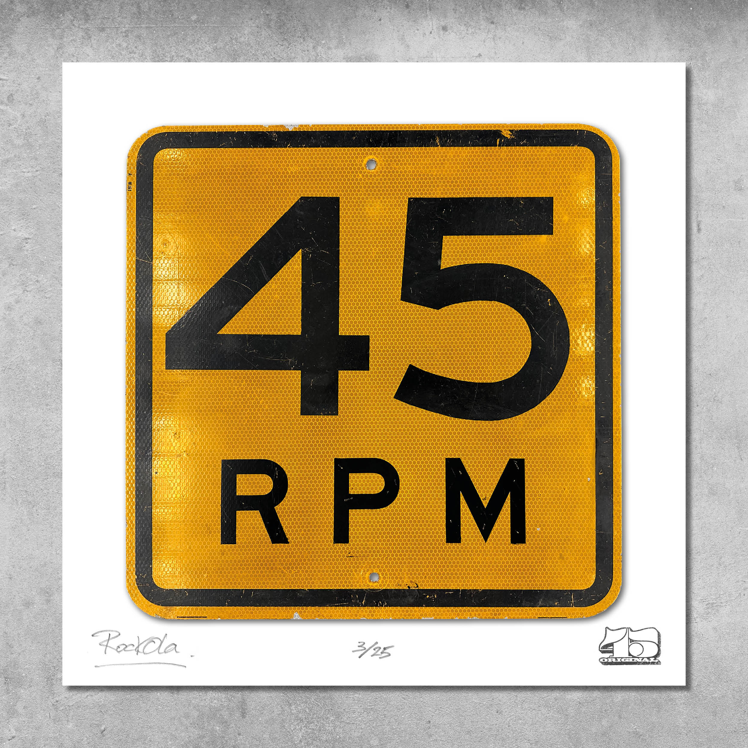 The RocknRoad Sign Print - 45 RPM US by My Store features a yellow and black metal-style sign with 45 RPM, inspired by vinyl records. Limited edition of 25, this unique print comes against a gray concrete background.