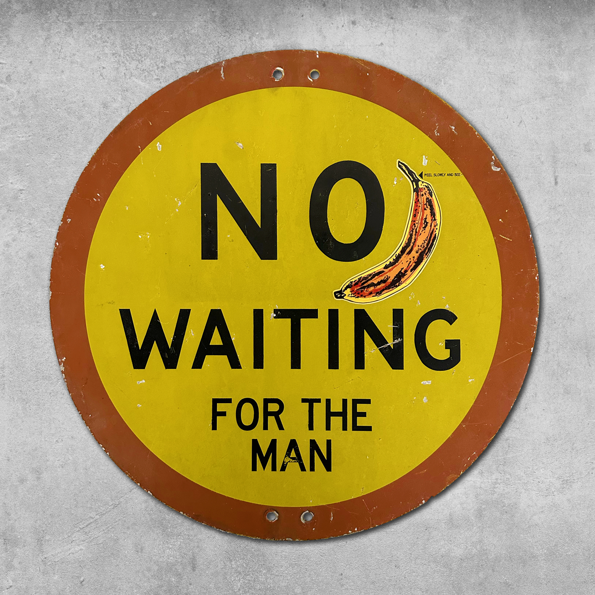 The 45 Original RocknRoad Sign - No Waiting For The Man is a round yellow UK metal sign with a brown border, Andy Warhol-style banana art, and is mounted on a gray textured wall.